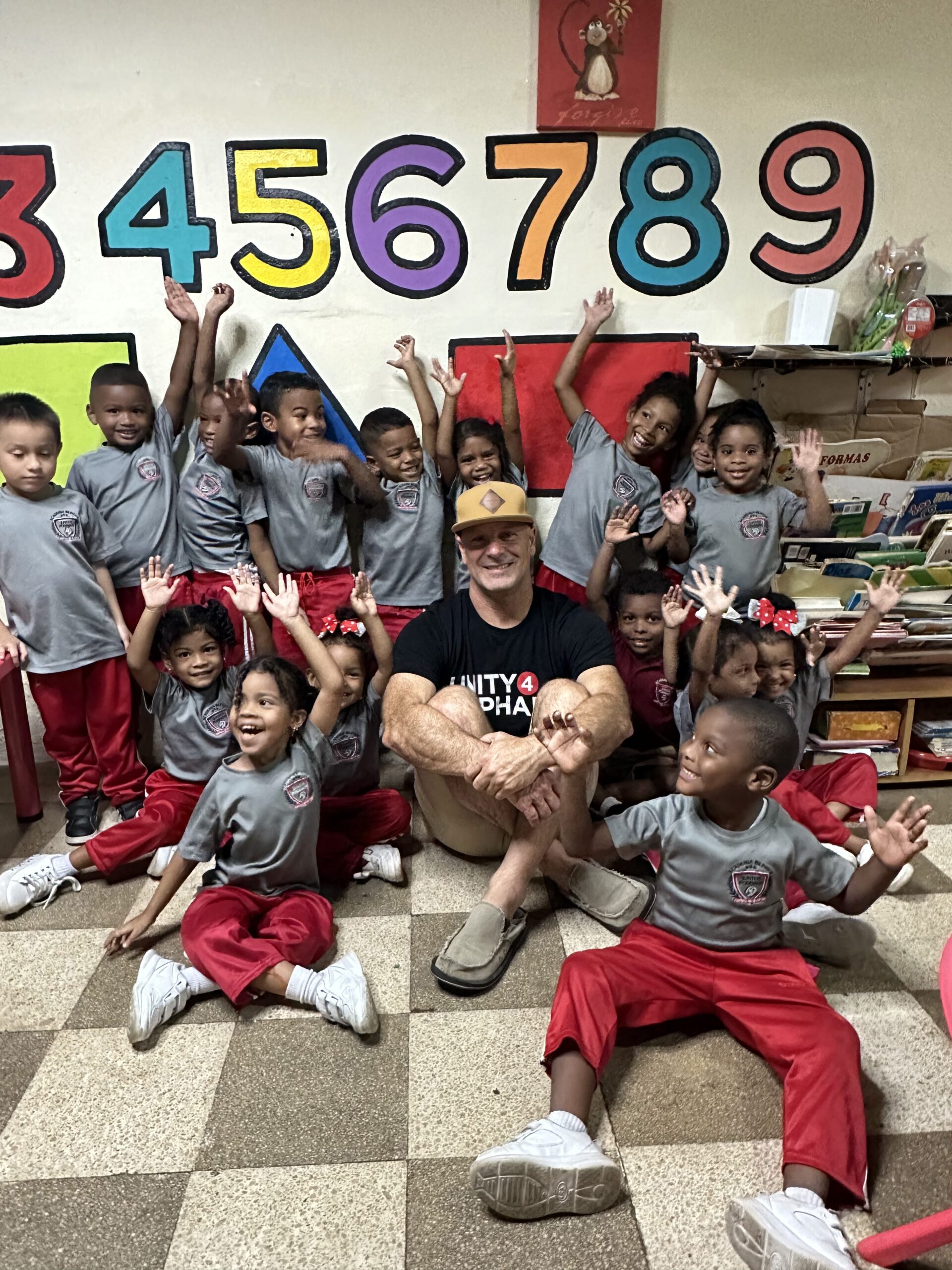A group of vulnerable children with their hands raised smiling excitedly while surrounding U4O founder Joe Brandi sitting on the floor smiling.